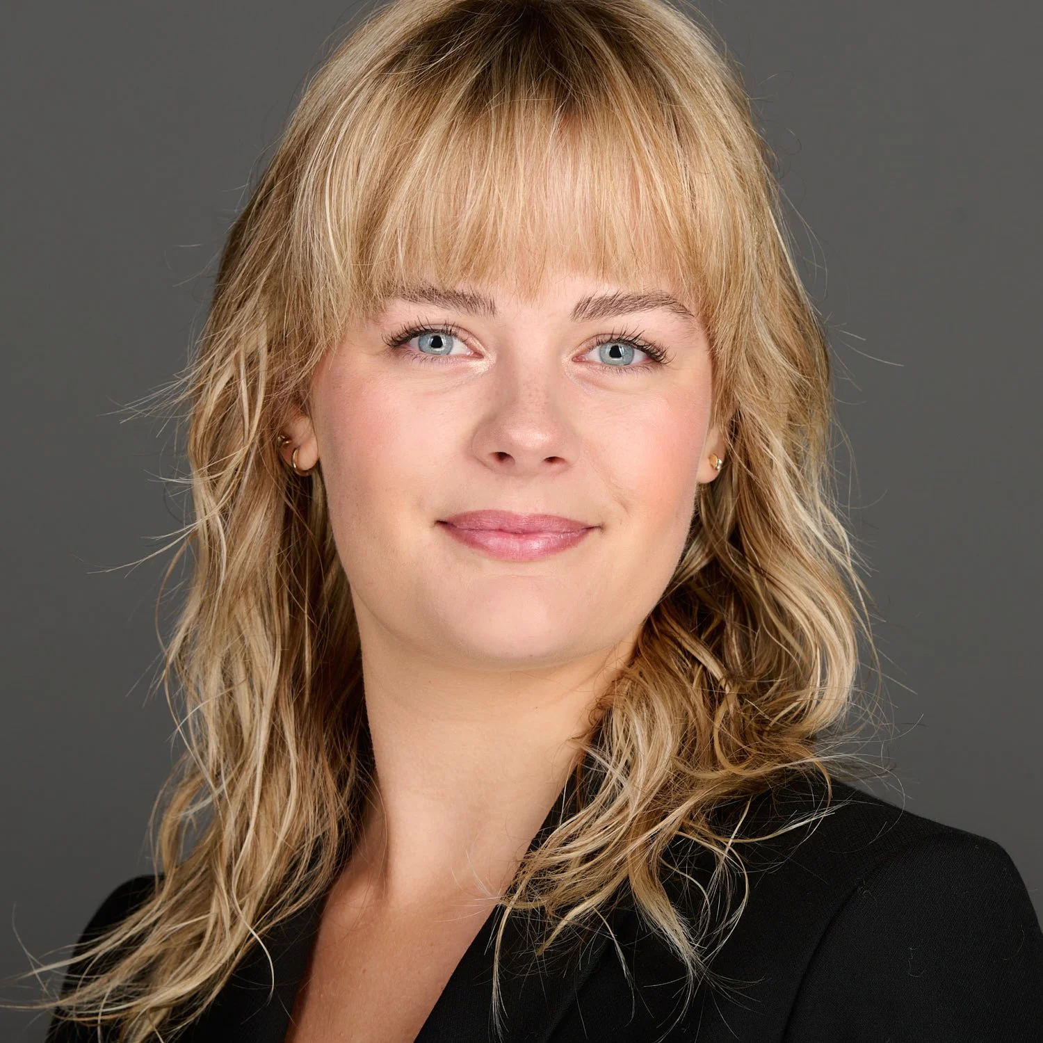 A woman's headshot with blonde, wavy hair, blue eyes, and a black blazer against a gray background.