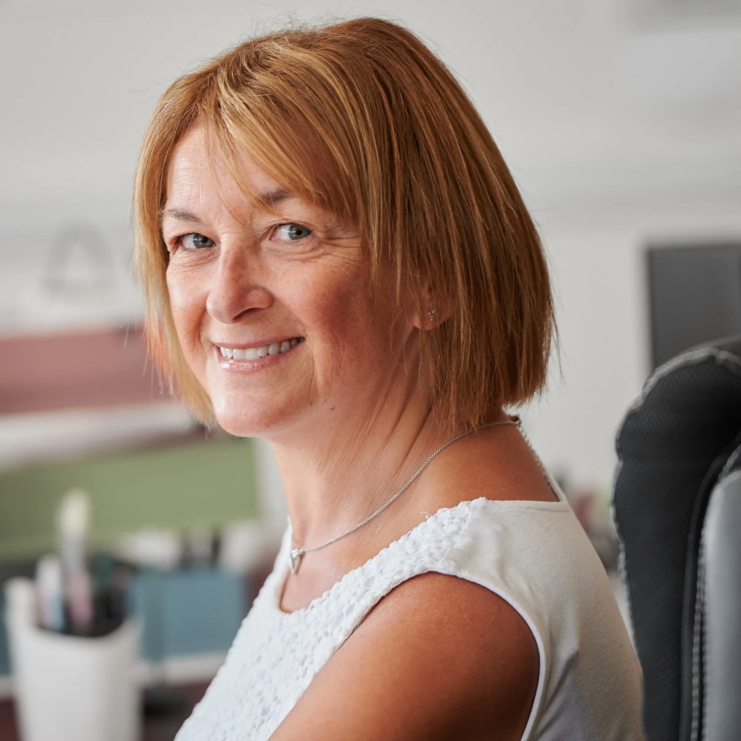 A middle-aged woman with shoulder-length red hair smiling, sitting in an office environment.