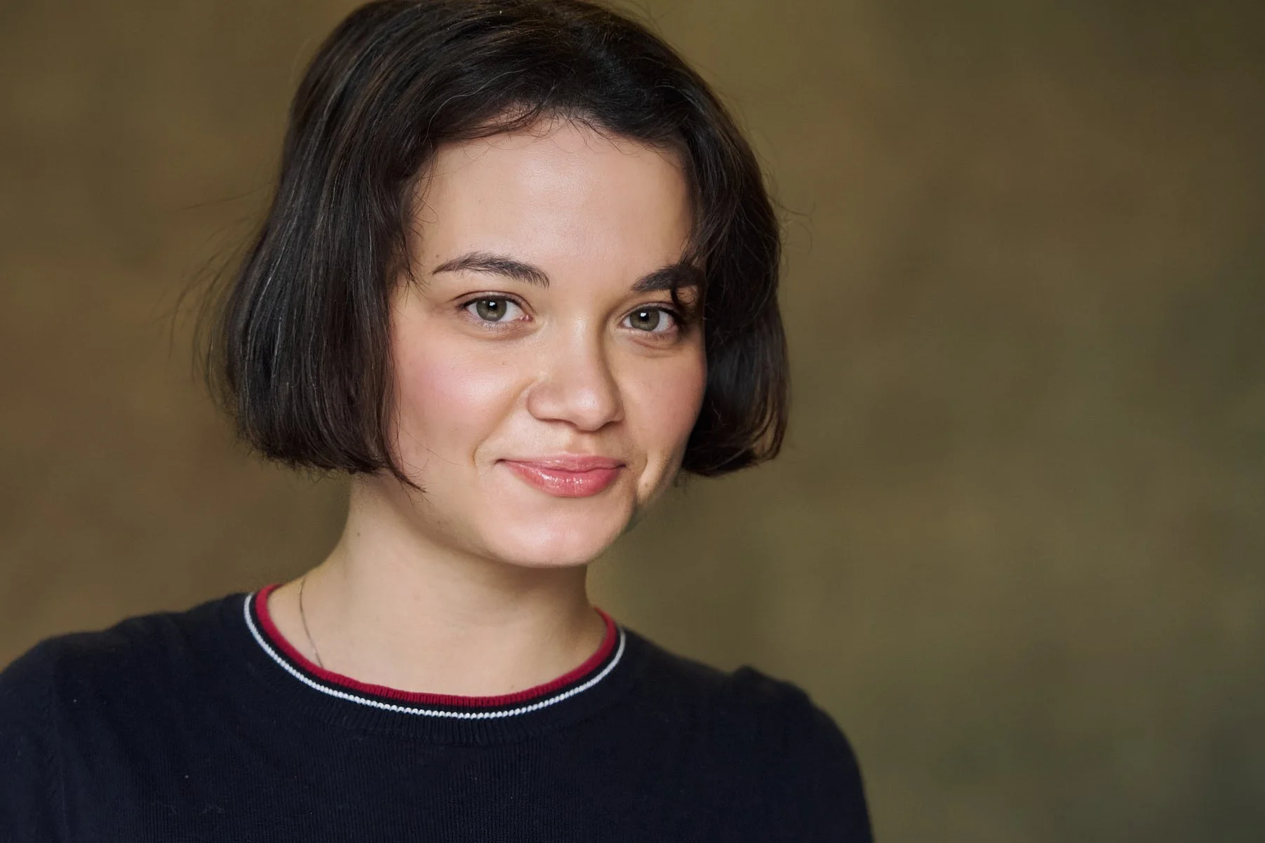 A young woman with short dark brown hair, fair skin, and blue eyes, smiling softly, wearing a black top with red, white, and blue trim around the collar, against a blurred brownish background.