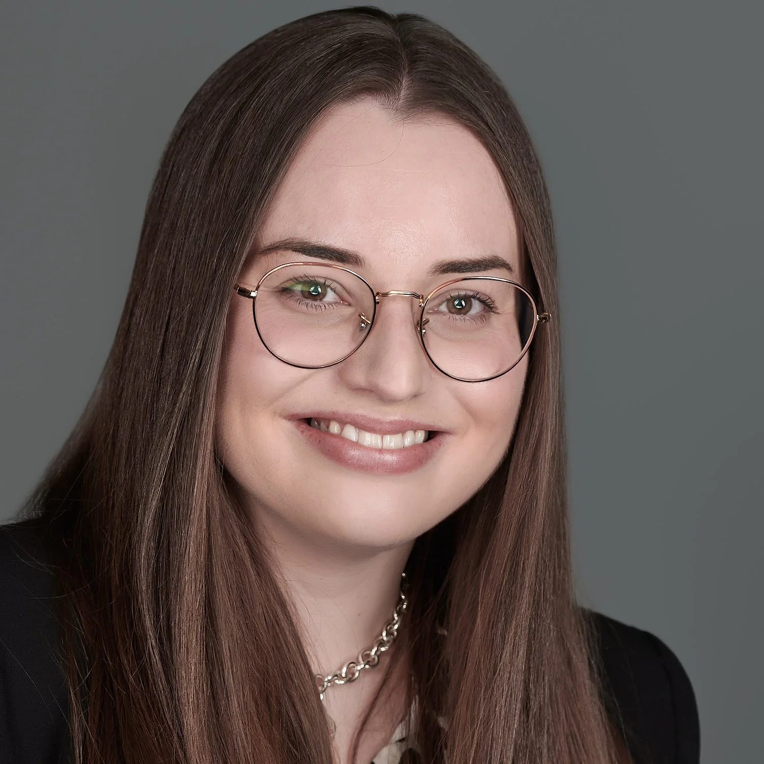 A young woman with long brown hair, wearing round glasses and a silver chain necklace, smiling at the camera against a plain gray background.