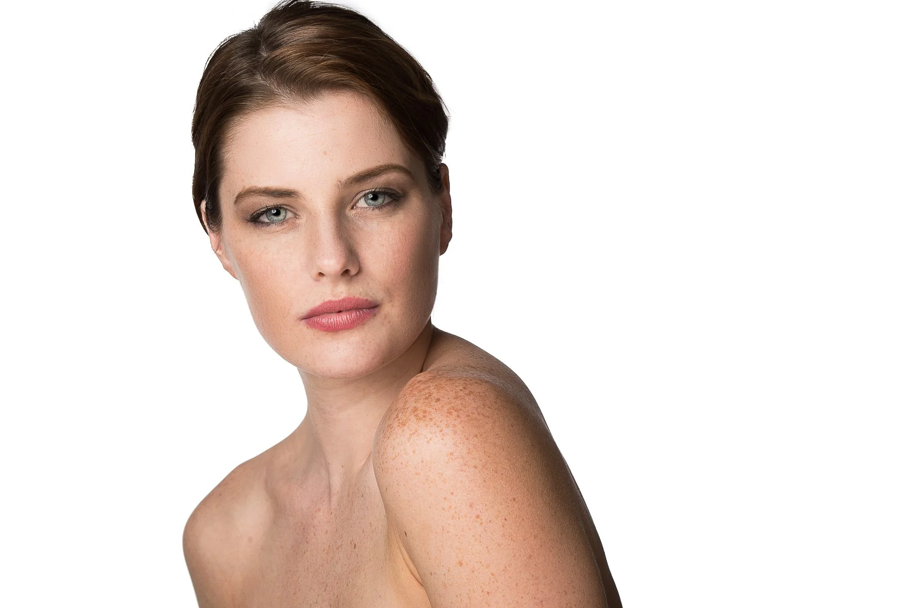 A portrait of a woman with light skin, short brown hair, and blue eyes, looking at the camera with a neutral expression, with her shoulder and upper arm visible and freckles on her shoulder.