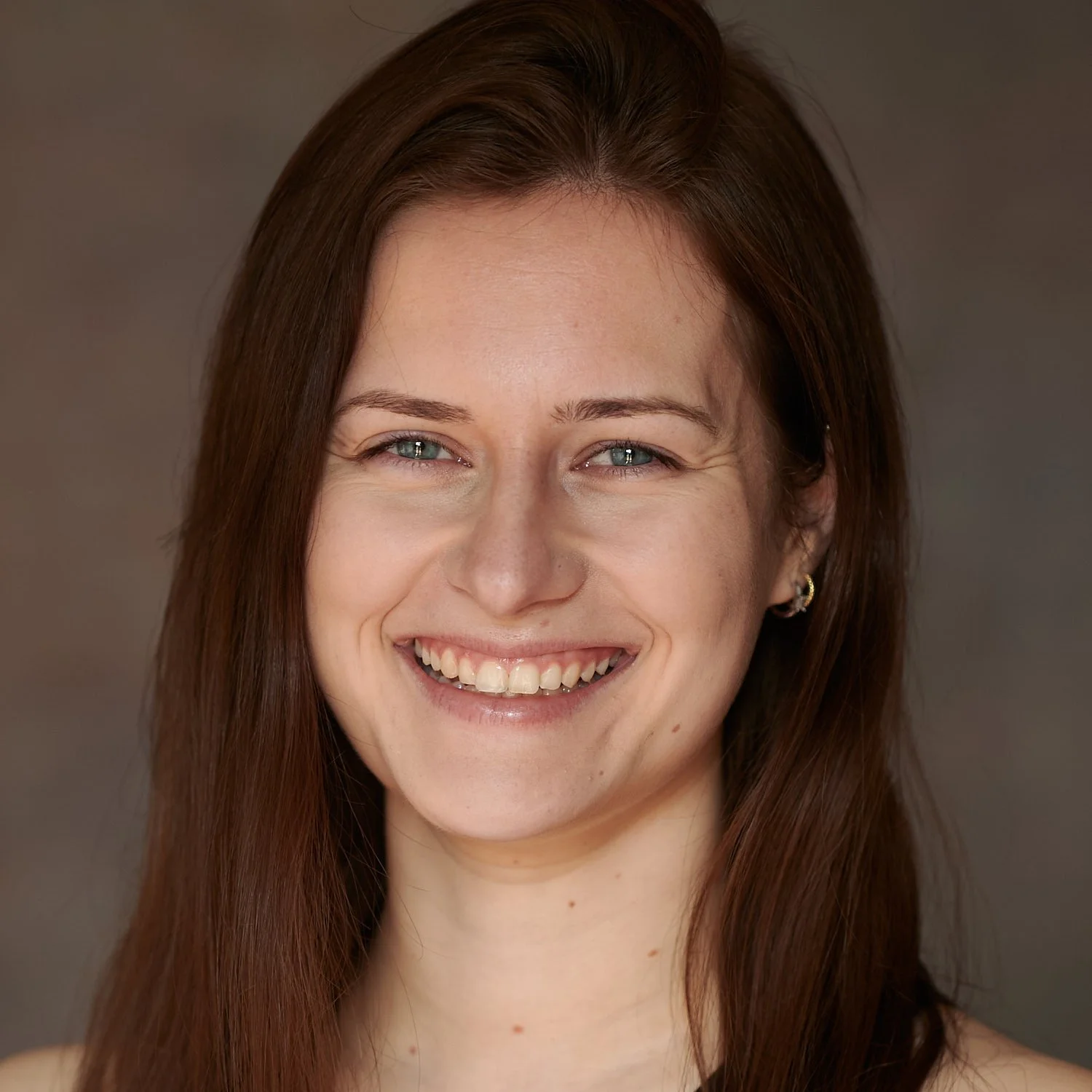 Close-up portrait of a woman smiling with blue eyes and long brown hair, wearing earrings, against a neutral background.