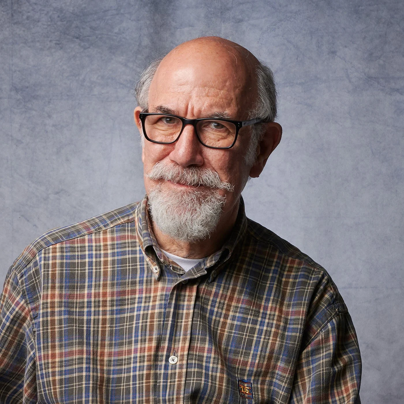 A portrait of an older man with glasses, a gray beard, and mustache, wearing a plaid shirt against a textured gray background.