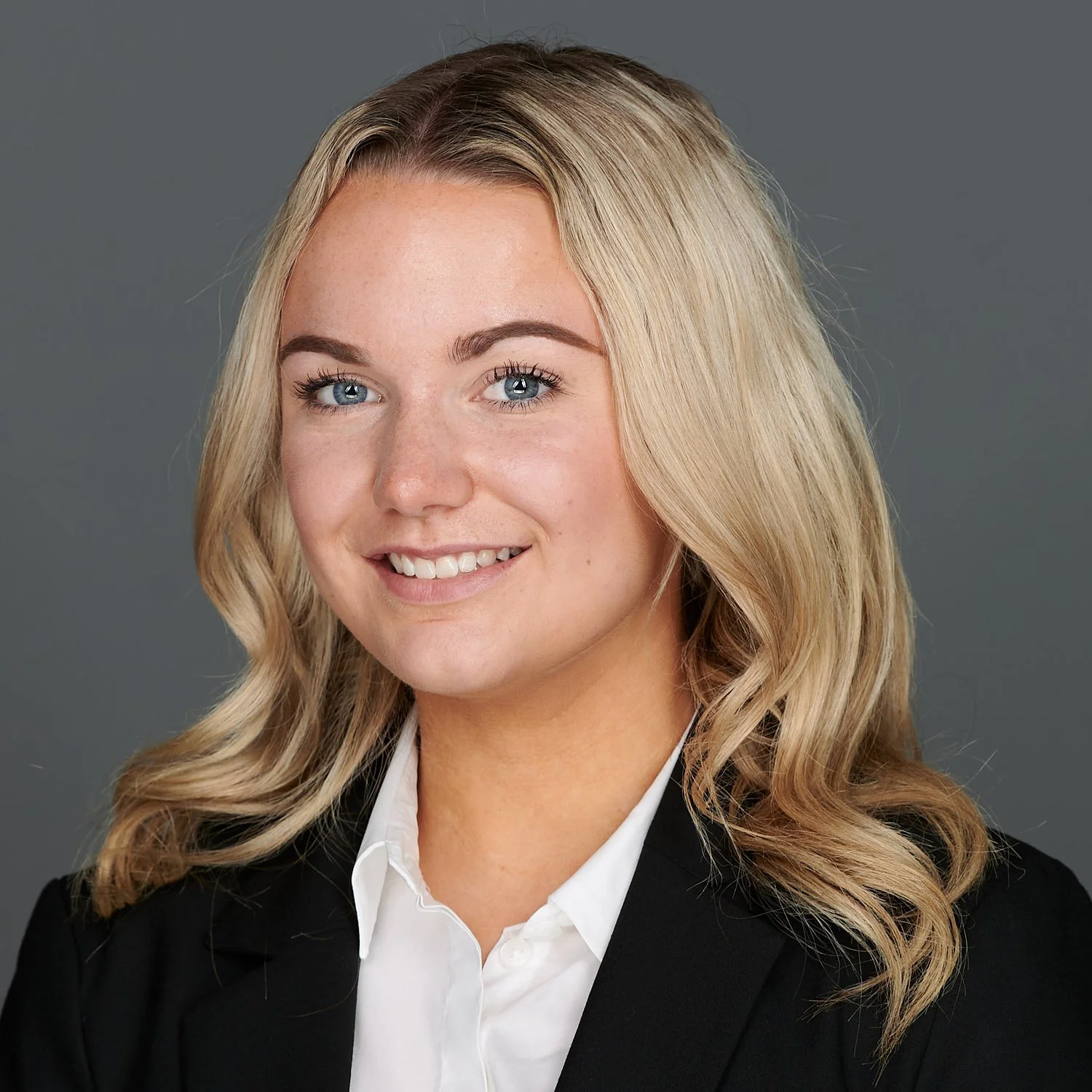 Professional headshot of a young woman with blonde hair, blue eyes, wearing a black blazer and white shirt, smiling against a gray background.