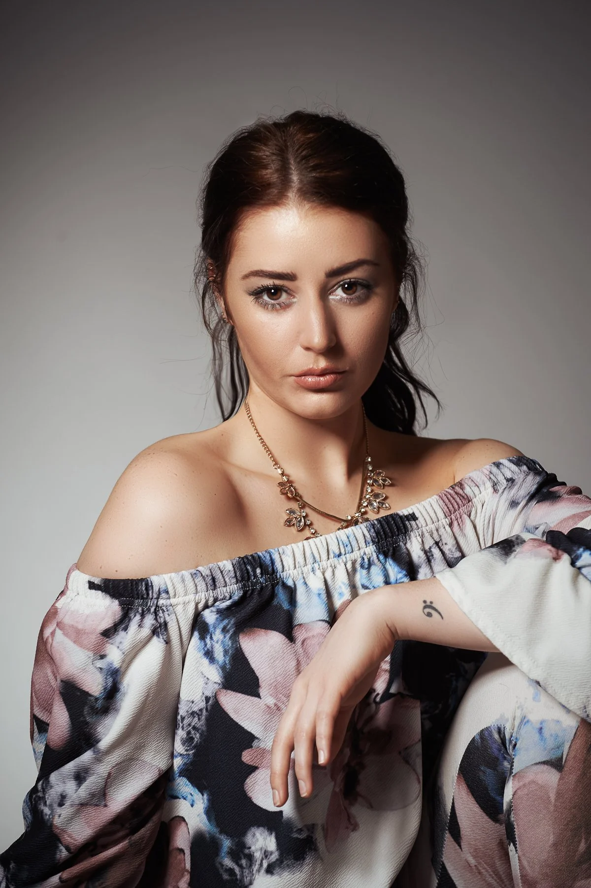 A young woman with long dark hair, wearing an off-shoulder floral top, a necklace, and displaying a small tattoo on her wrist, posing against a neutral gray background.