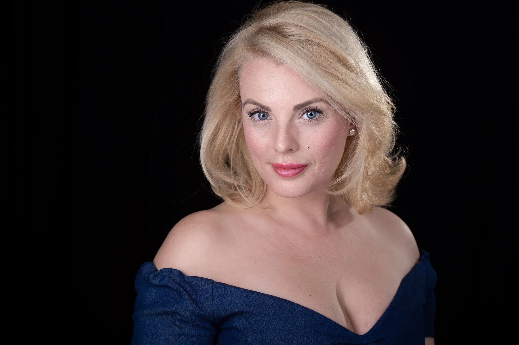 Blonde woman with blue eyes, wearing a dark off-the-shoulder top, against a black background.