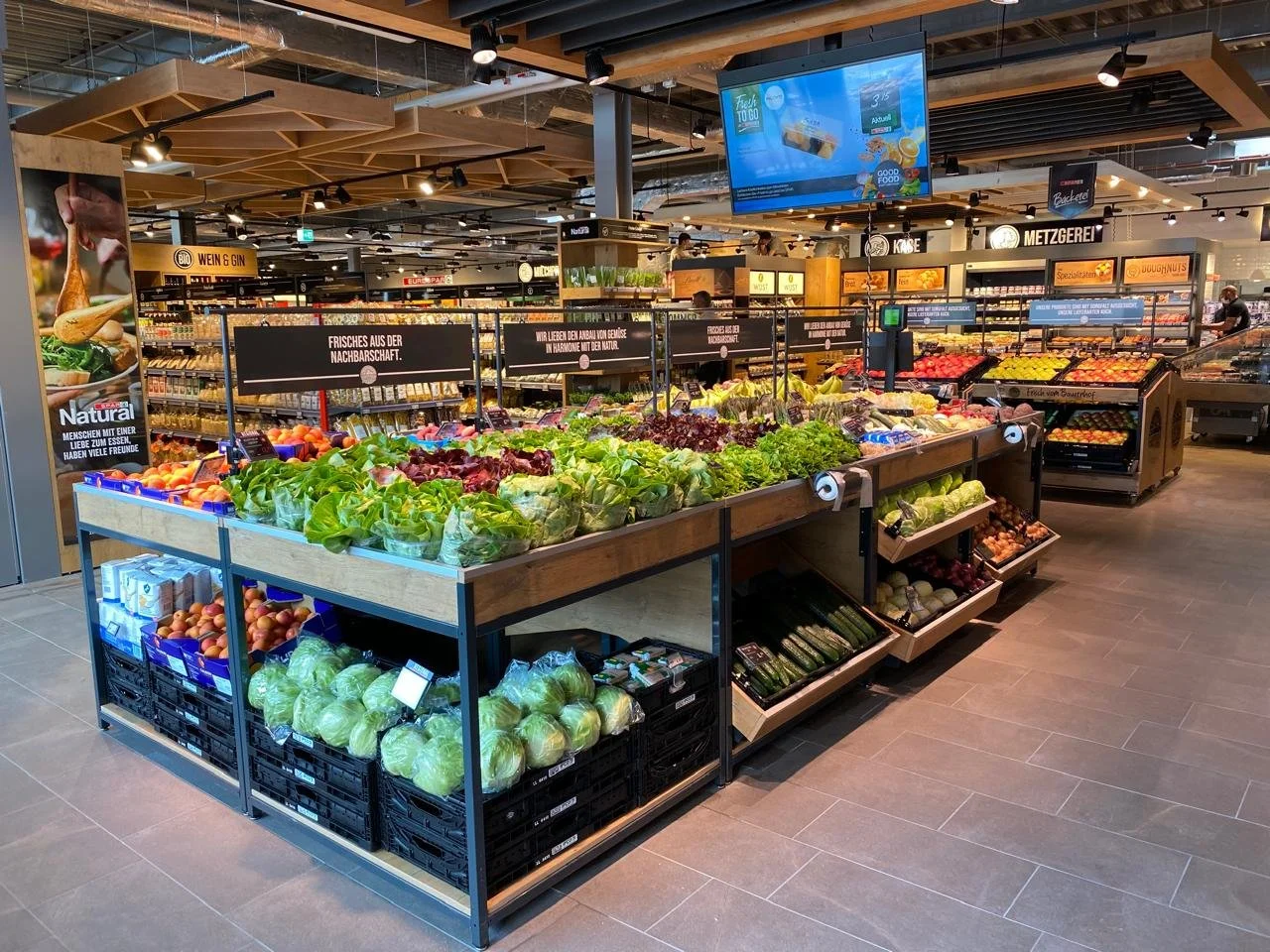Fresh produce section in a grocery store with lettuce, cabbage, onions, and other vegetables on display, with signage and a digital screen overhead.