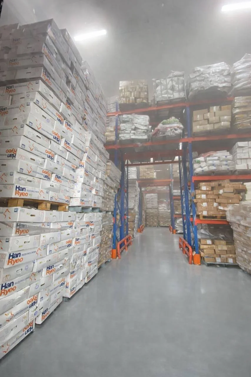 Warehouse aisle with shelves stacked with boxes and pallets, some wrapped in plastic, with a clear concrete floor.