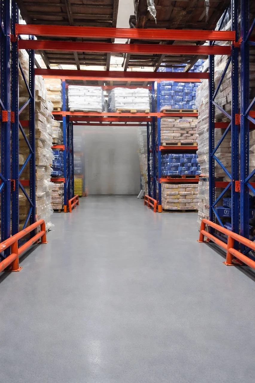 Empty industrial warehouse aisle with tall blue and orange metal shelving units stocked with stacks of construction materials, including bricks and tiles.