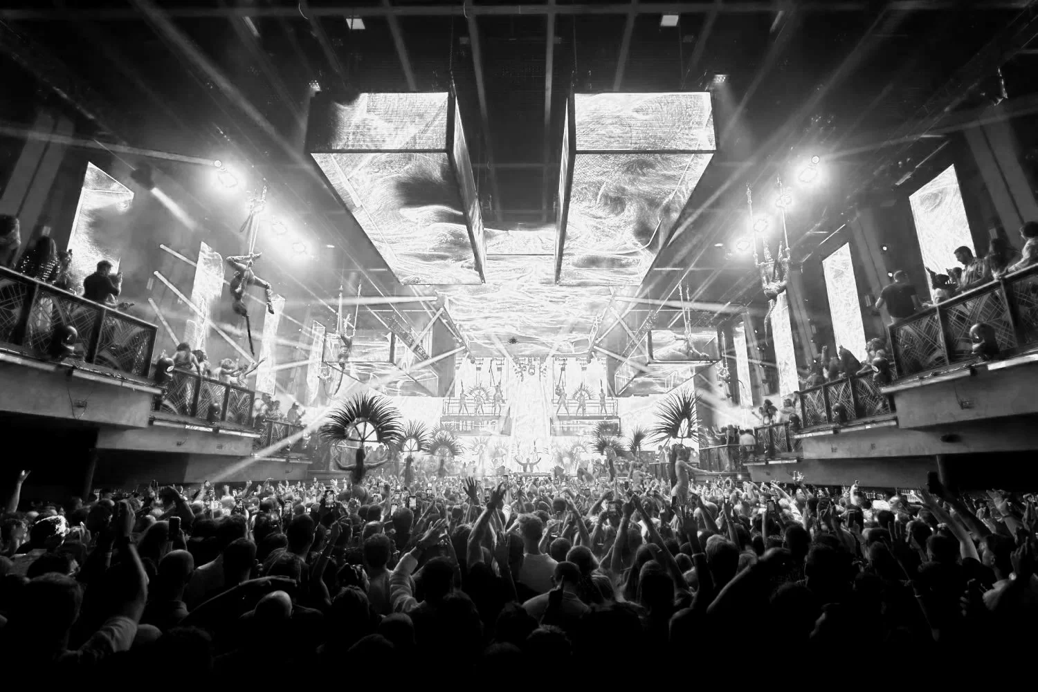 Black and white photo of a crowded indoor concert or club with a large stage, dancers, and hanging aerial performers, with many audience members raising their hands.