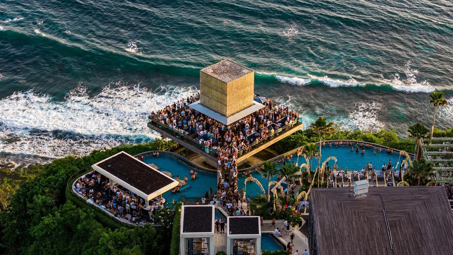 Aerial view of a crowded rooftop pool party near the ocean, with multiple people gathered around pools and on a large deck, featuring lush green trees and buildings nearby.
