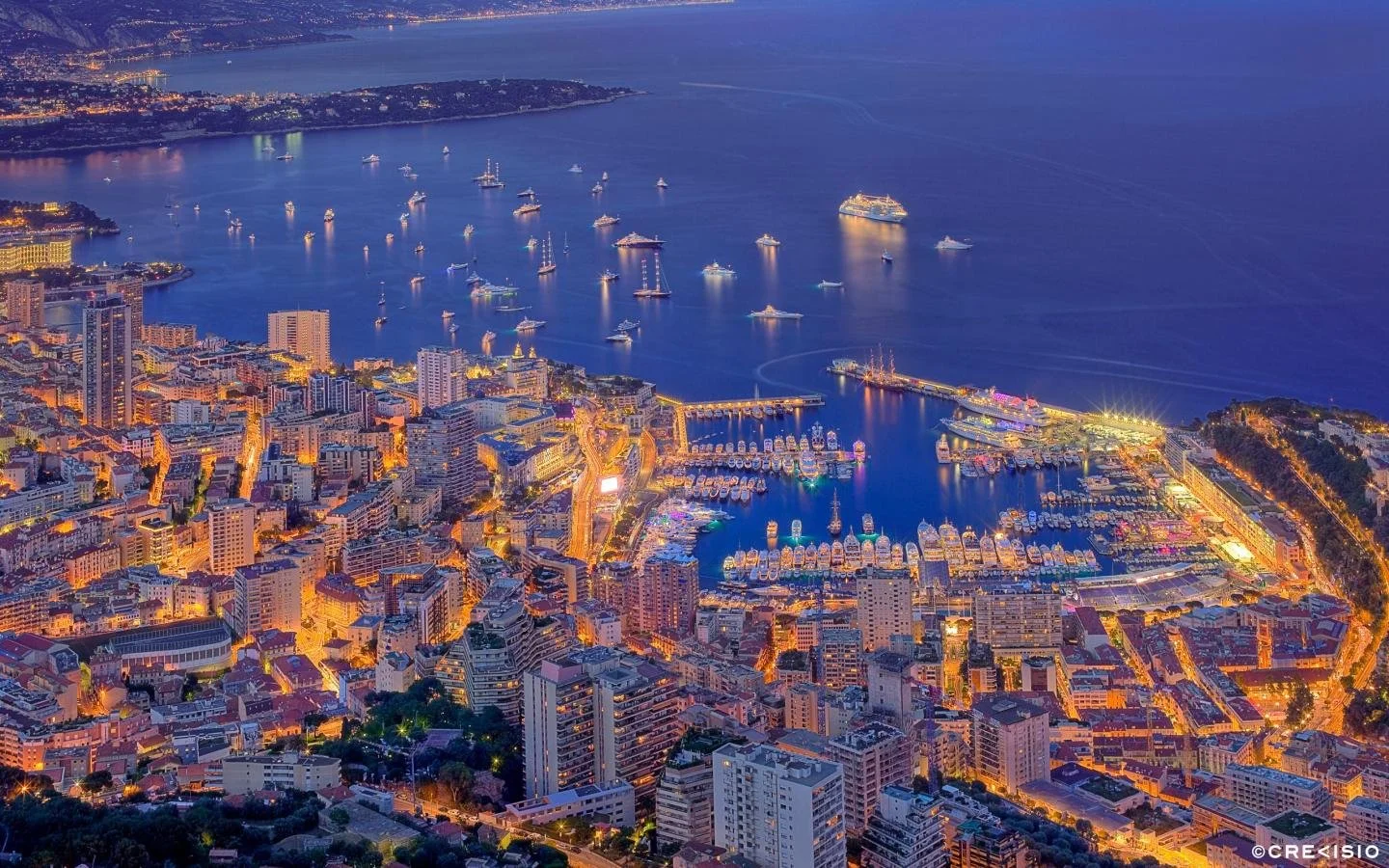 An aerial view of a city at dusk with numerous buildings, a marina with boats, and a large body of water with cruise ships and yachts.