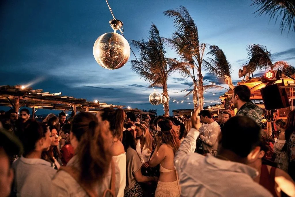 People dancing and socializing at an outdoor party with palm trees, string lights, and disco balls hanging from the sky during dusk.