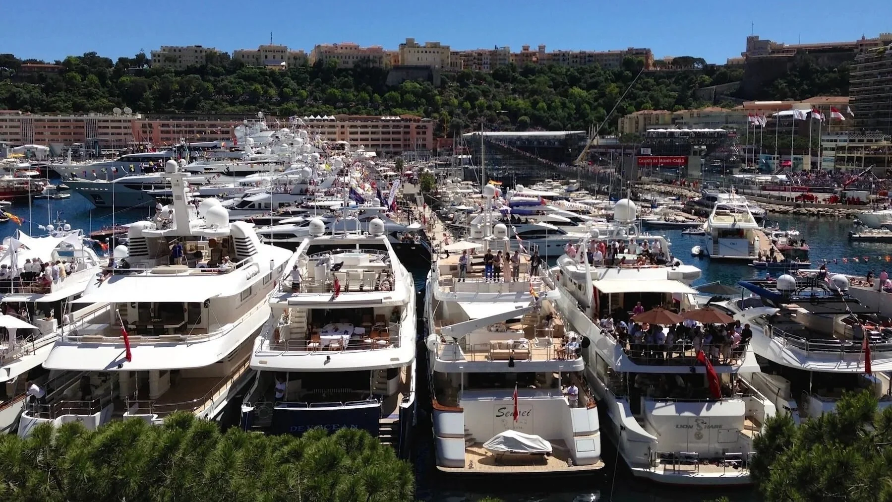 A harbor filled with numerous luxury yachts docked side by side, with people enjoying on the decks and walking along the docks. In the background, there are buildings on a hillside and a clear blue sky.