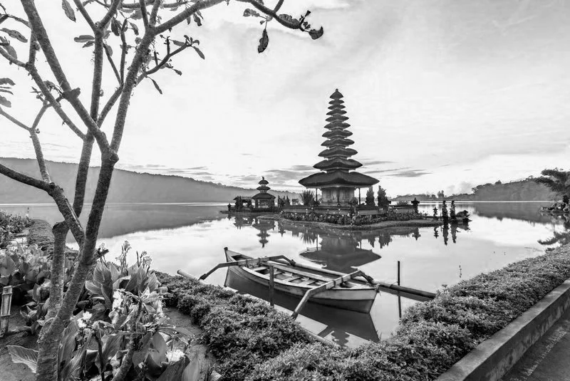 A black and white photo of a peaceful lake with a pagoda-style structure on the water, surrounded by a low wall and lush vegetation. A boat is docked near the shore, and hills are visible in the background.