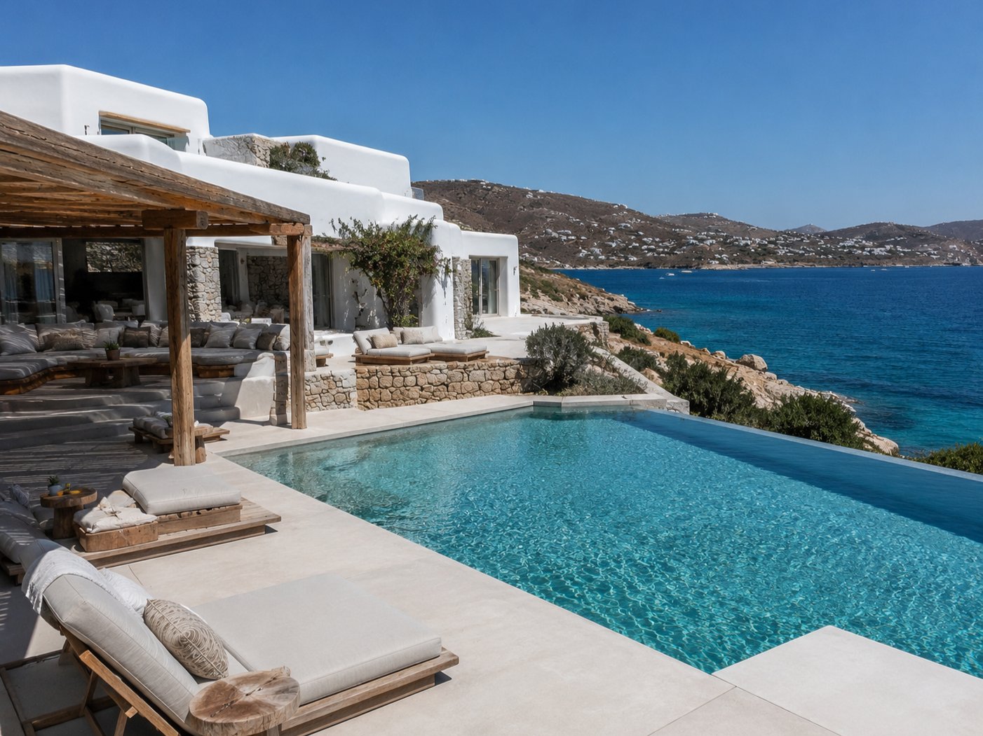 Luxury villa with a swimming pool overlooking the ocean, Mediterranean-style white building, outdoor lounge area with cushions, wooden pergola, scenic coastal landscape in Greece.