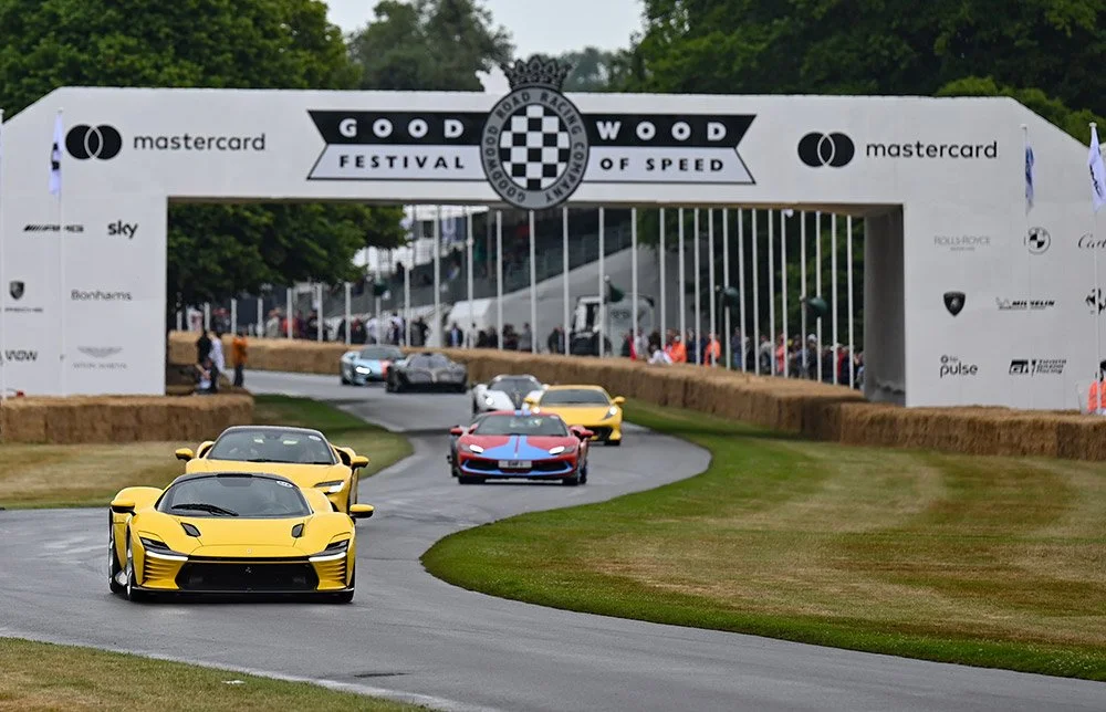 Several sports cars, including a yellow McLaren and a red Ferrari, racing on a track during the Goodwood Festival of Speed, with banners and spectators in the background.