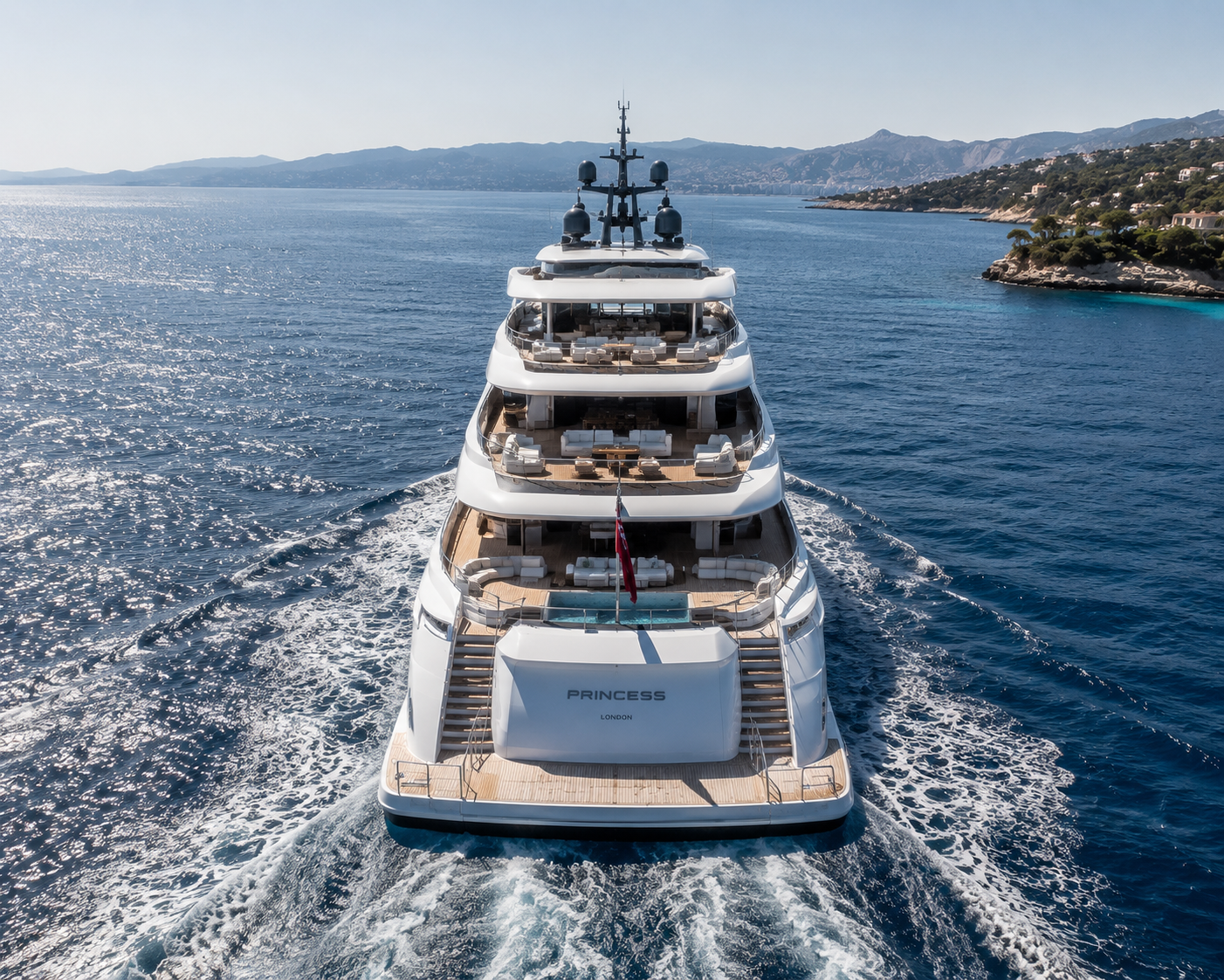 A large luxury yacht named Princess sailing on the ocean with a deck featuring outdoor seating and a swimming pool, and a coastline with mountains and houses in the background.