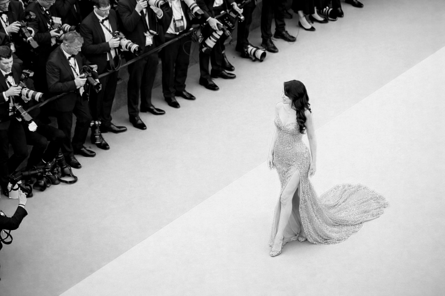 A woman in a sparkling gown with a thigh-high slit walking on a fashion runway, surrounded by photographers taking pictures.