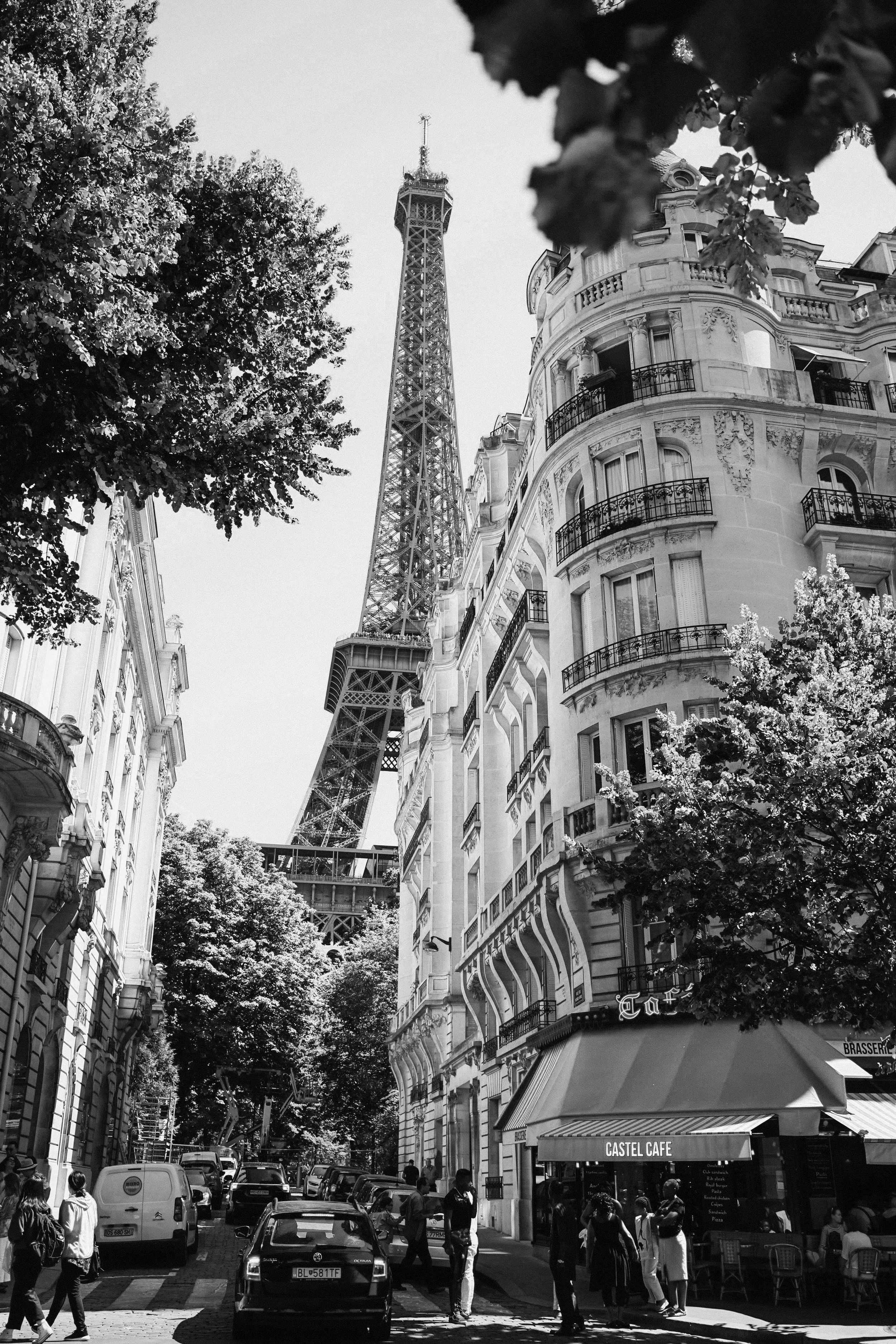 Street scene in Paris with the Eiffel Tower in the background, pedestrians crossing, cars parked, a café named Castel Café, and historic buildings.