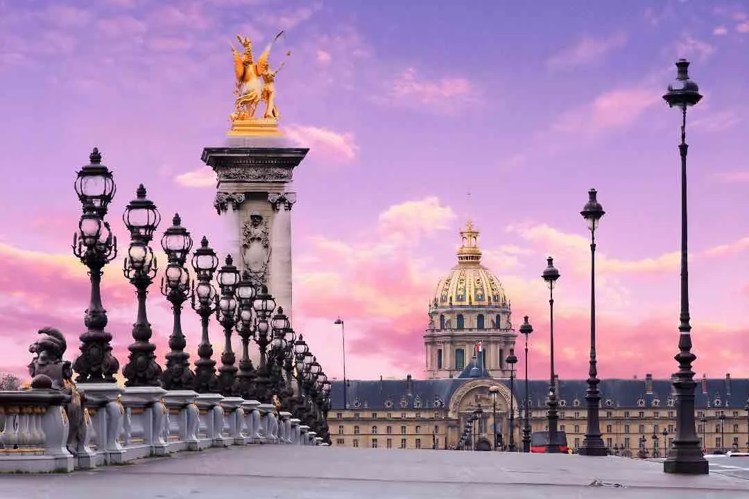View of the Pont Alexandre III bridge in Paris, featuring ornate lamp posts and a golden statue of a winged horse on a column, with a domed building in the background during sunset.