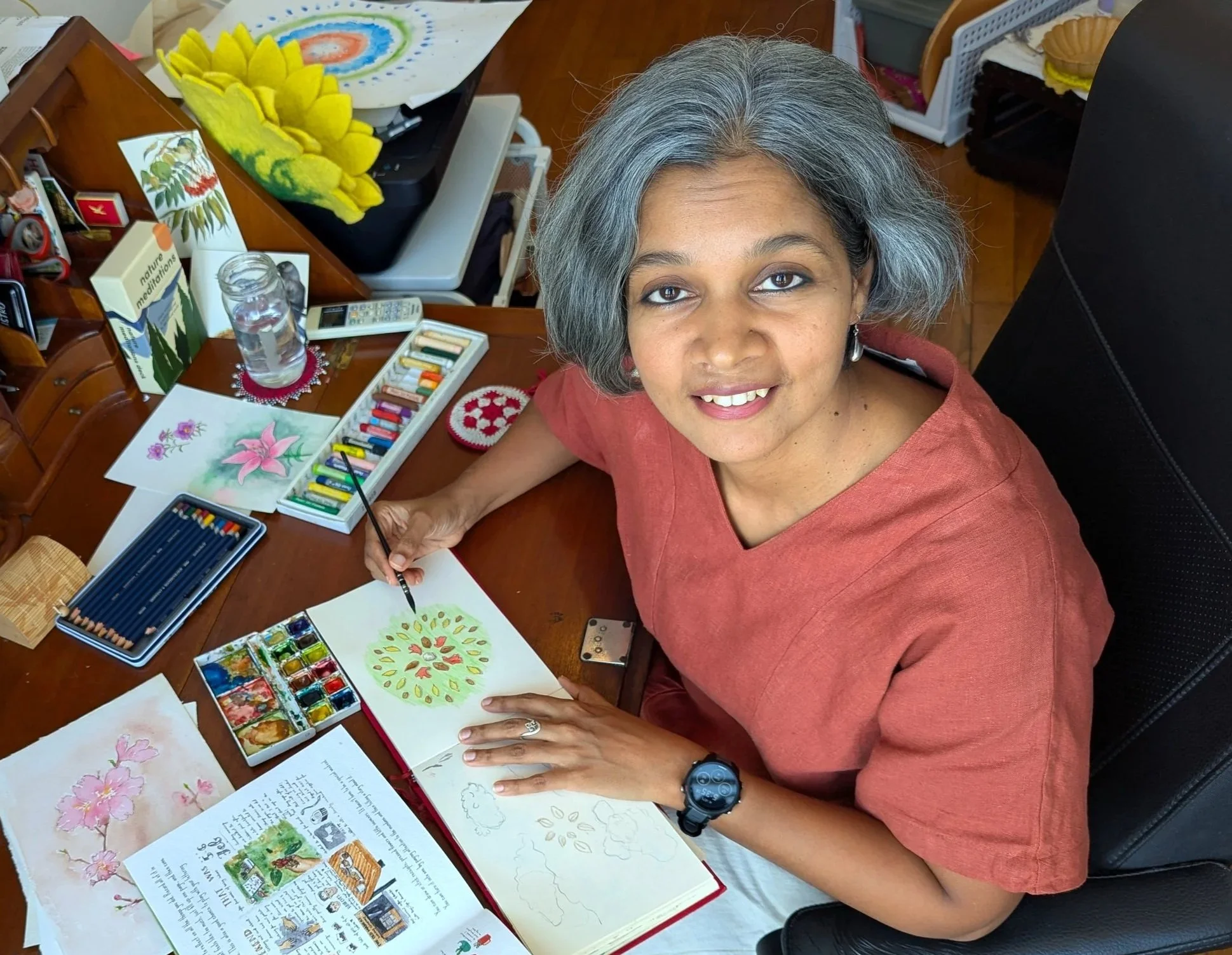 A woman with gray hair and a brown top sitting at a desk, painting botanical illustrations with watercolors, surrounded by art supplies, books, and decorative items in a home office.