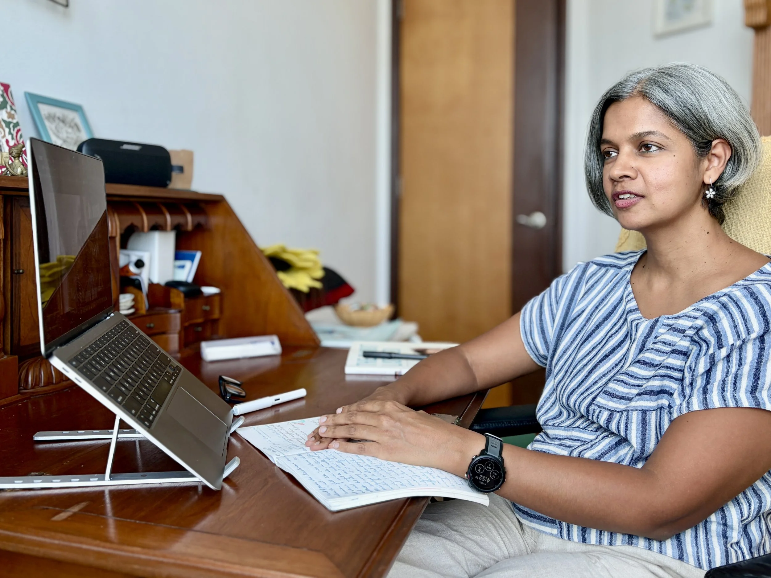A woman with gray hair and earrings is sitting at a wooden desk, looking at a laptop on a stand, with papers, a pen, and a smartwatch on her wrist.