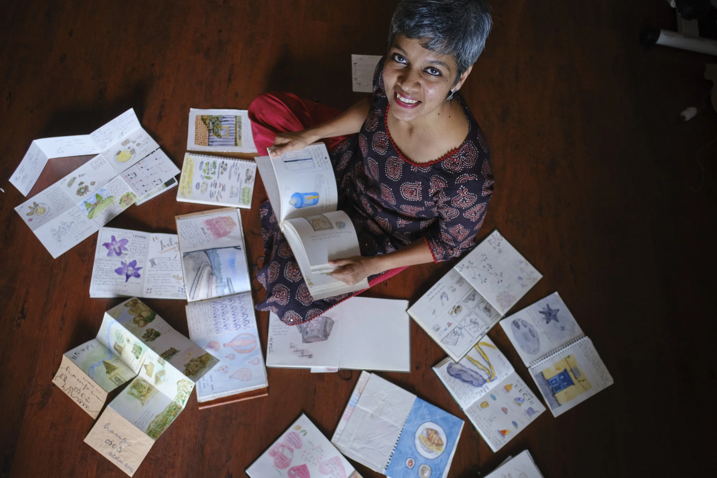 A woman with short gray hair sitting cross-legged on a wooden floor, surrounded by open sketchbooks and notebooks filled with colorful drawings and illustrations, looking up at the camera and smiling.