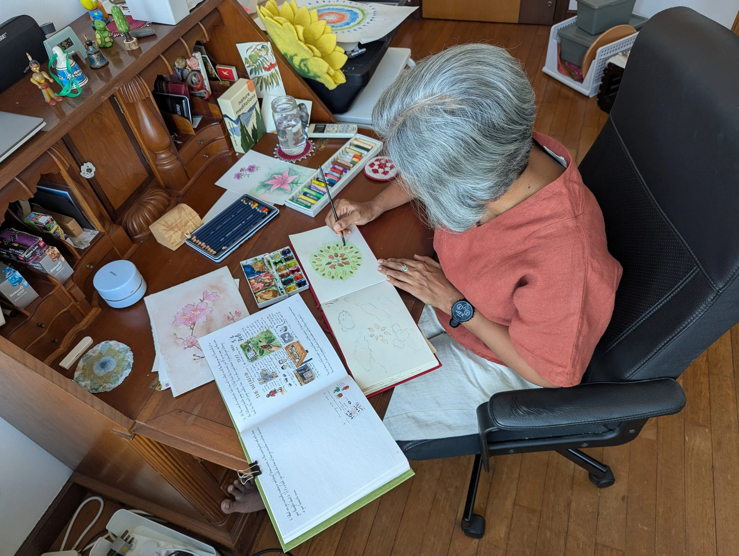 A person sitting at a wooden desk, painting watercolor art with various paint supplies around, including watercolor paints, brushes, and artwork. The desk has personal items and decorations, including small figurines, a jar of water, and colorful artwork.