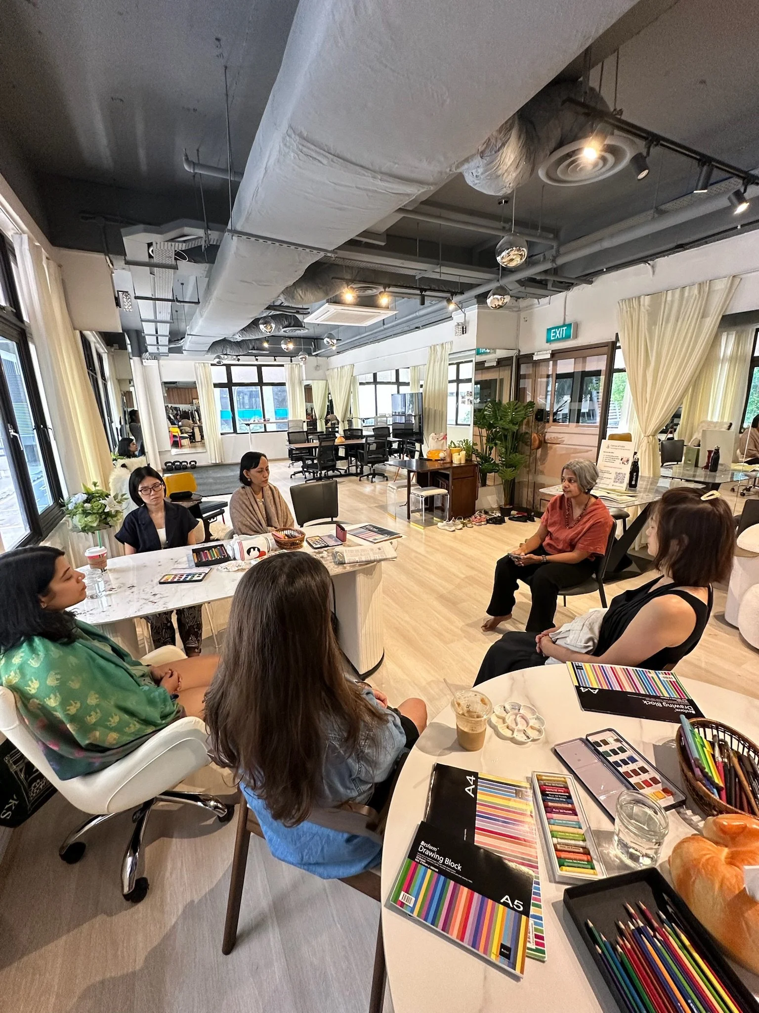 Group of women sitting in a circle in a spacious, well-lit room with large windows, engaged in a discussion or workshop, with art supplies and notebooks on the tables.