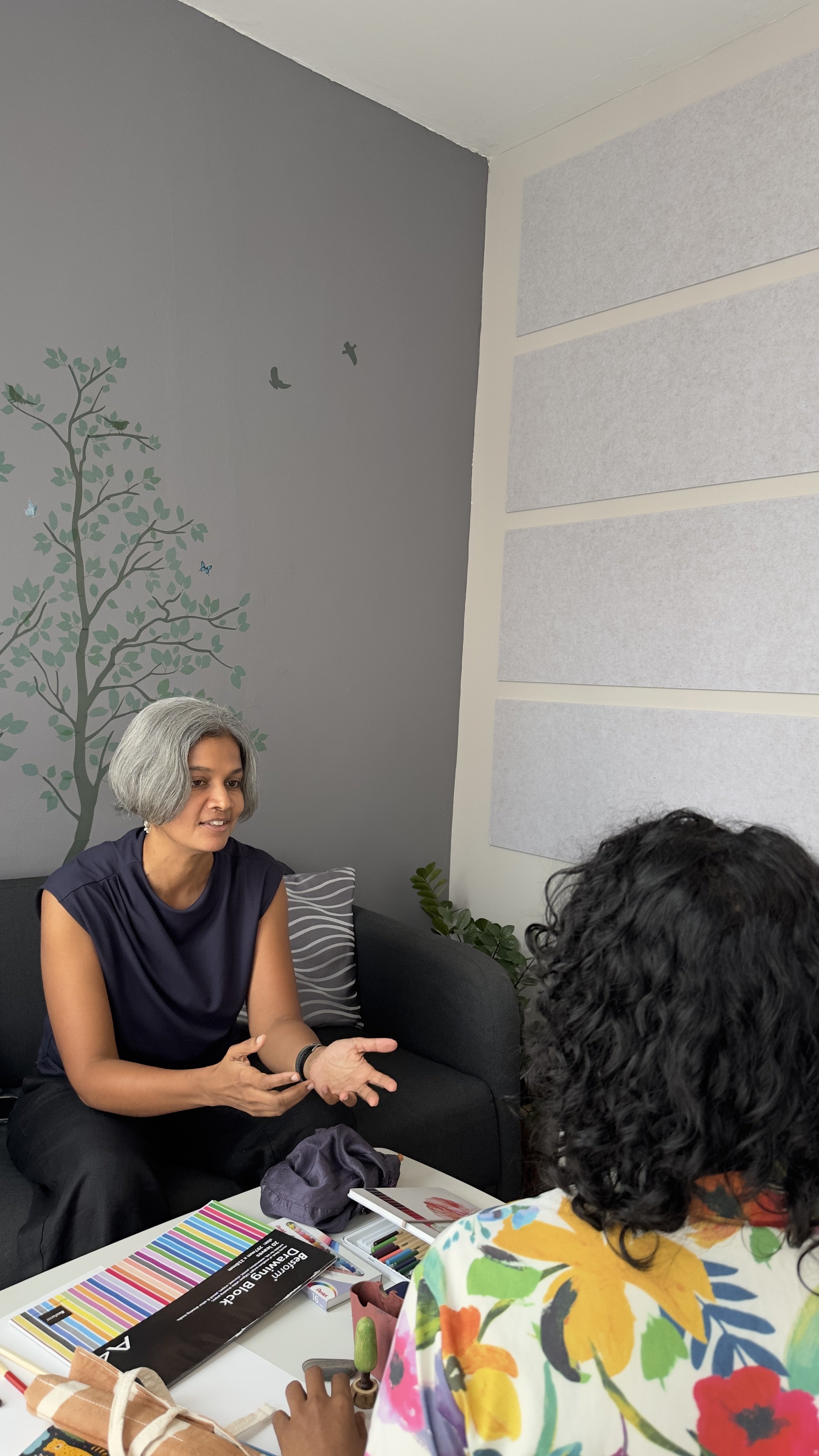 Two women having a conversation in a cozy room. One woman with short gray hair is gesturing with her hands, while the other woman has curly black hair and is wearing a colorful floral shirt.