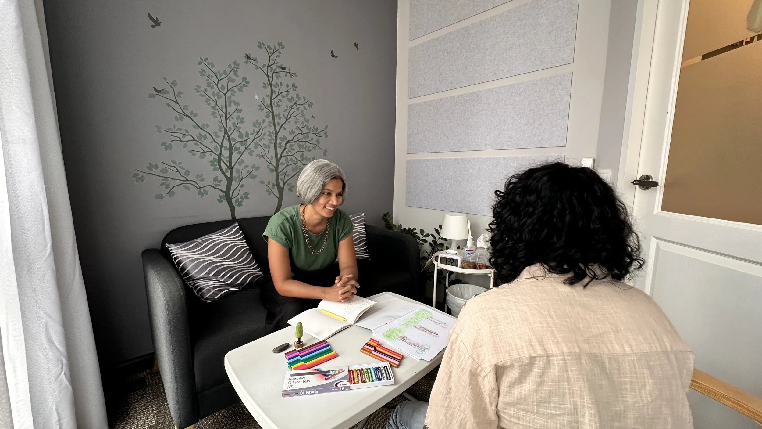 Two women sitting at a small white table with art supplies, including oil pastels and a drawing of trees, in a room with gray walls and a black couch.