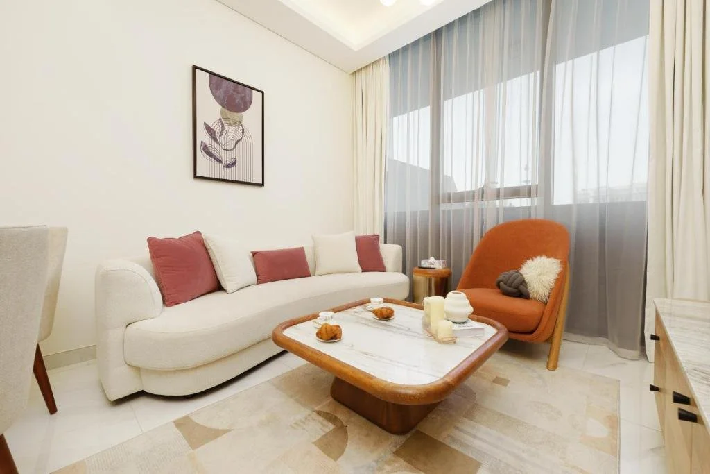Living room with a white sofa, pink and white pillows, an orange chair with a beige pillow, a marble-topped coffee table with candles and snacks, a small side table, art on the wall, and large windows with sheer and blackout curtains.