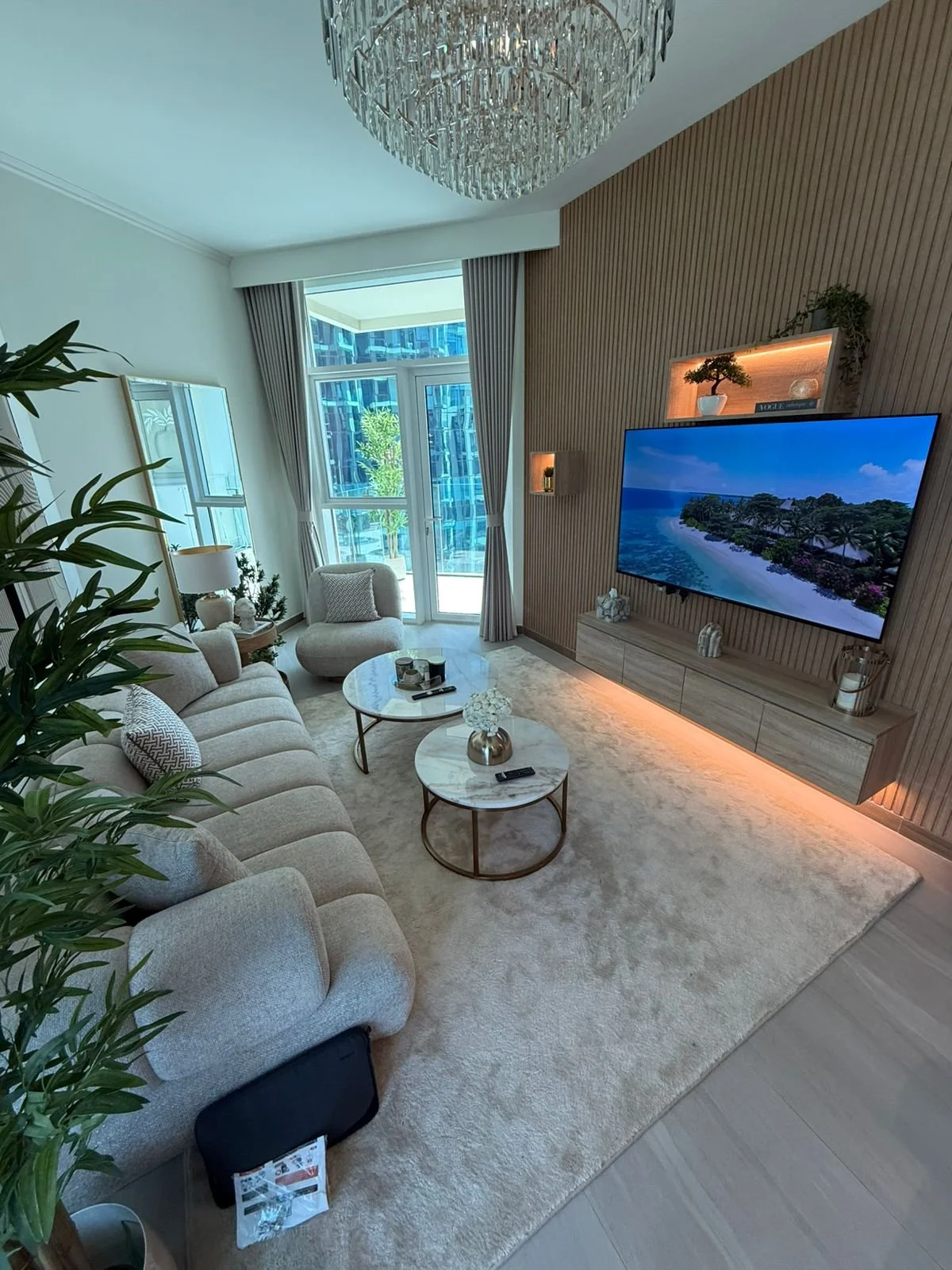 Modern living room with beige sofa, armchair, marble coffee tables, flat-screen TV, wood-paneled accent wall, and large balcony door with curtains, decorated with plants and decorative objects.