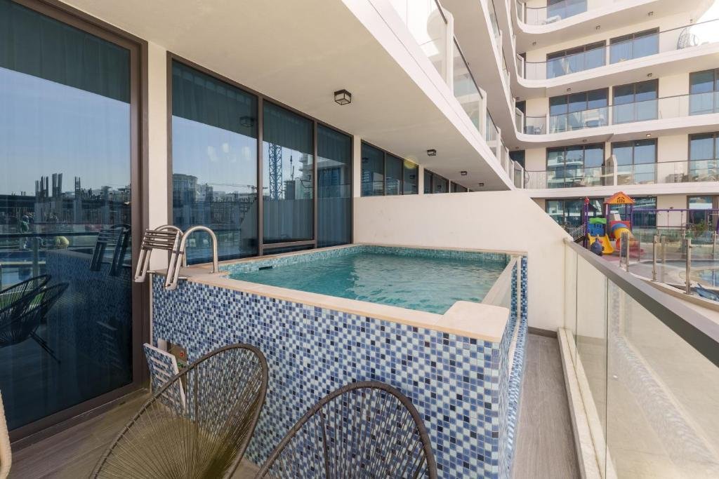 Balcony view of a small outdoor pool with mosaic tiles, overlooking a residential complex with glass balconies and a playground in the background.