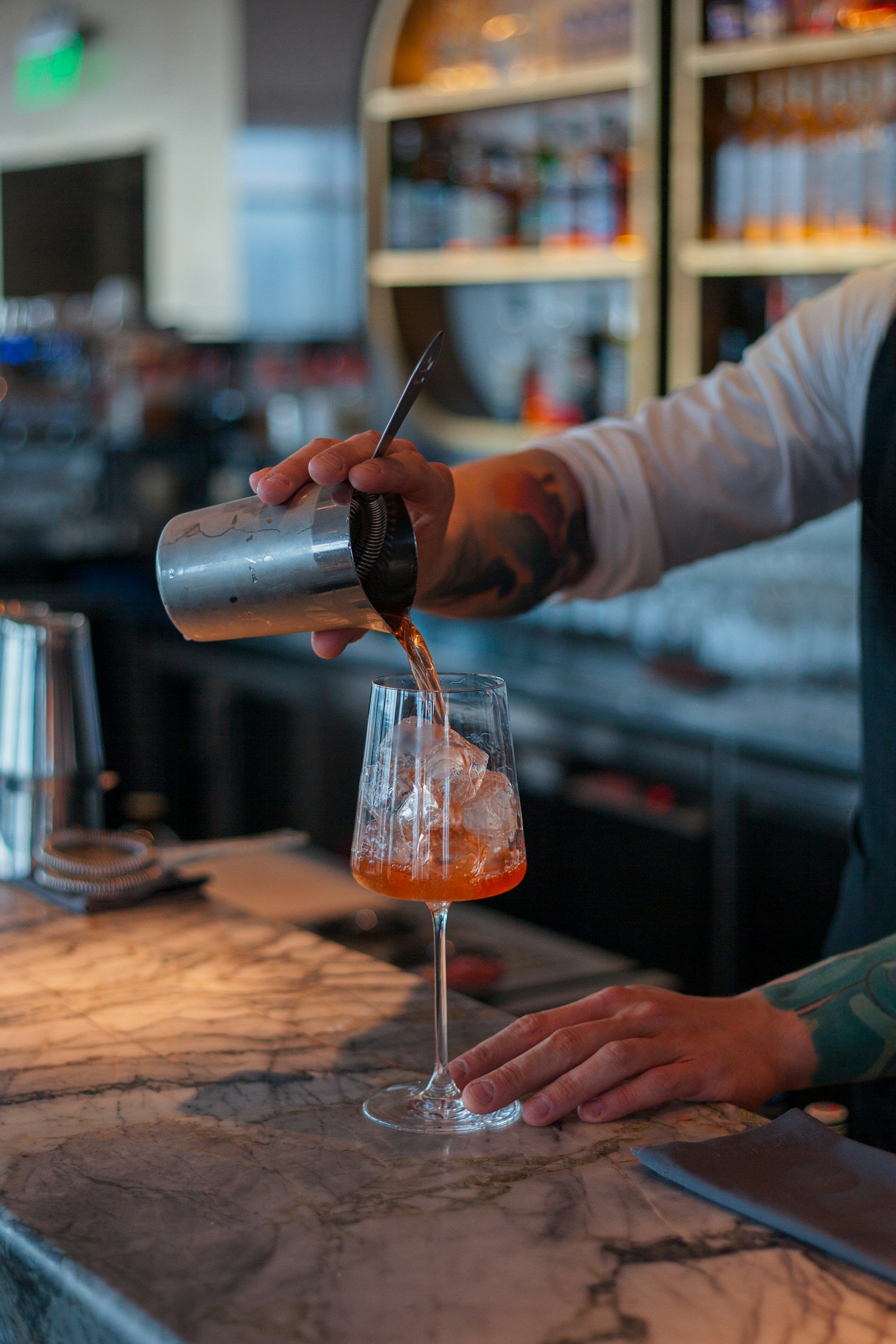 A bartender pours a dark cocktail over ice into a tall, curved glass with a long stem at a bar.