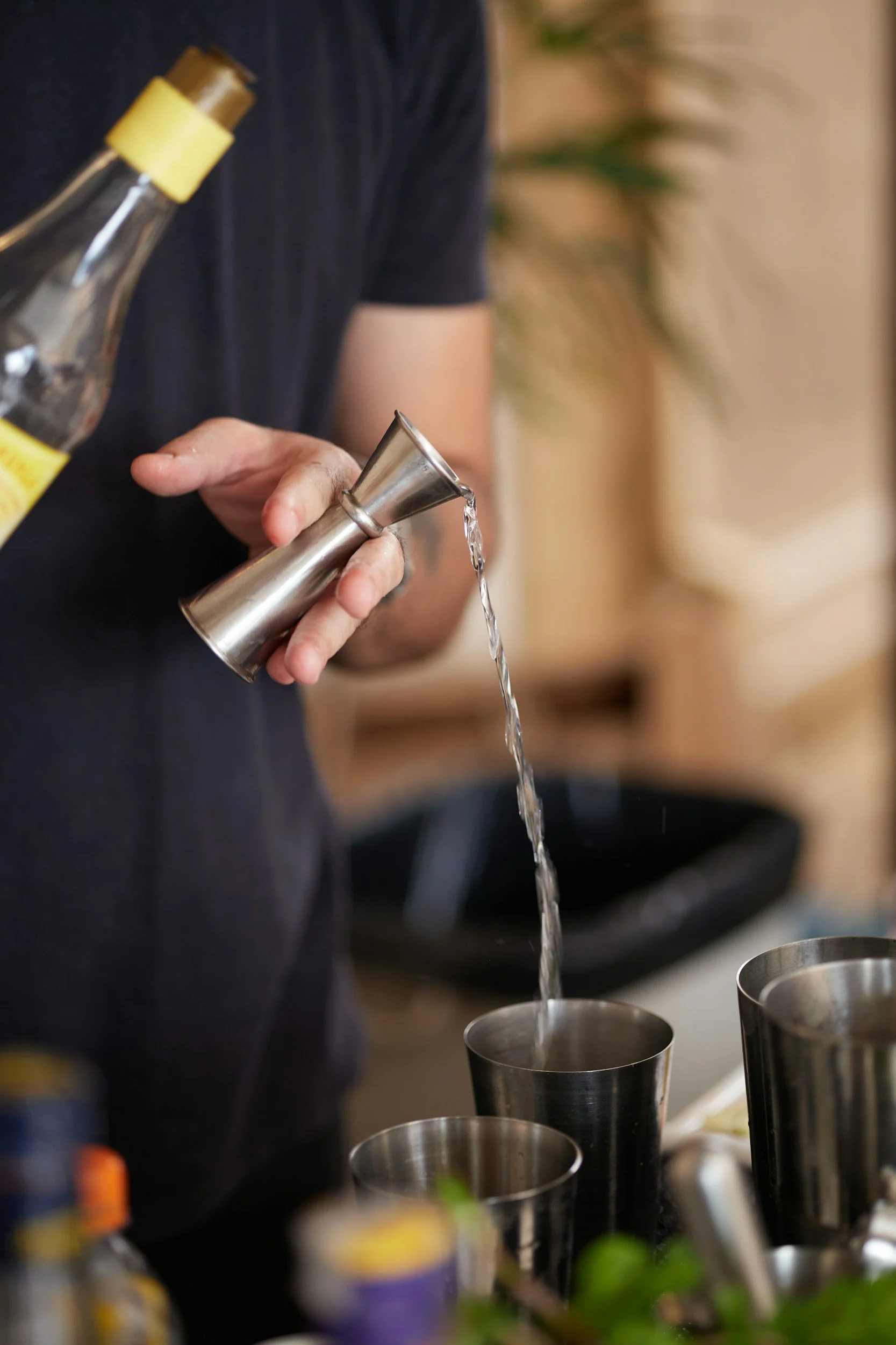 A person in a black shirt pouring a liquid from a jigger into a metal shaker in a bar or kitchen setting.