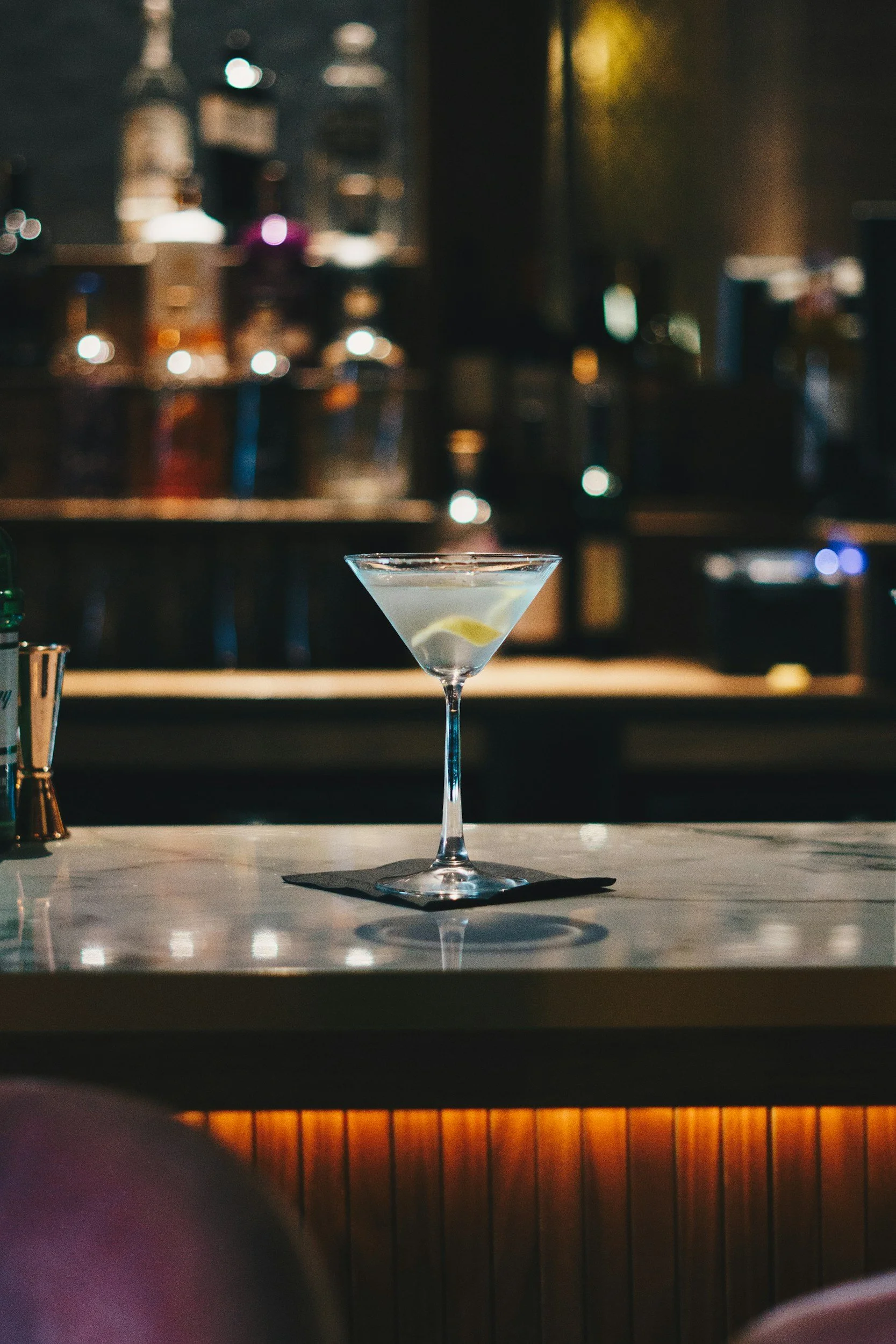 A martini glass filled with a clear cocktail, garnished with a lemon wedge, on a bar counter with a blurred background of bottles and bar equipment.