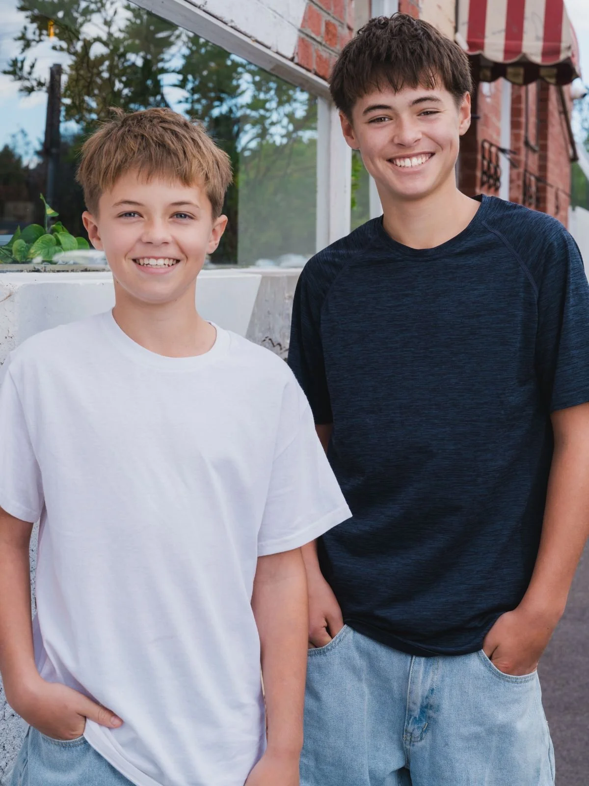 Two teenage boys with short brown hair smiling outdoors, standing in front of a brick building and a glass window, with trees in the background.