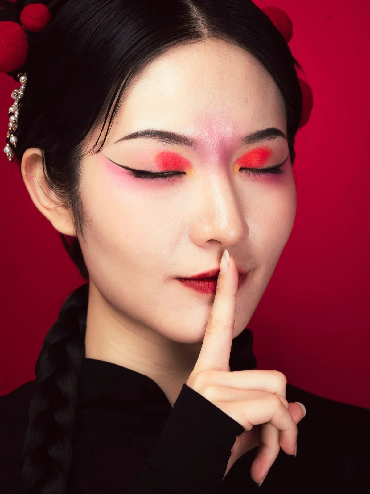 A woman with floral hair accessories and bold eye makeup, holding her finger to her lips in a shushing gesture against a red background.