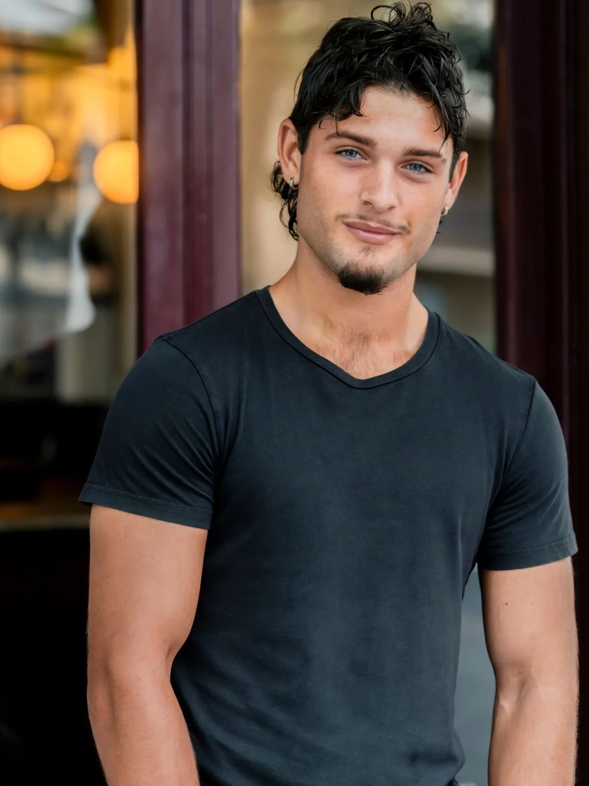 A young man with dark, curly hair and blue eyes is smiling, wearing a black t-shirt, standing outdoors near a building with glass windows and purple curtains.