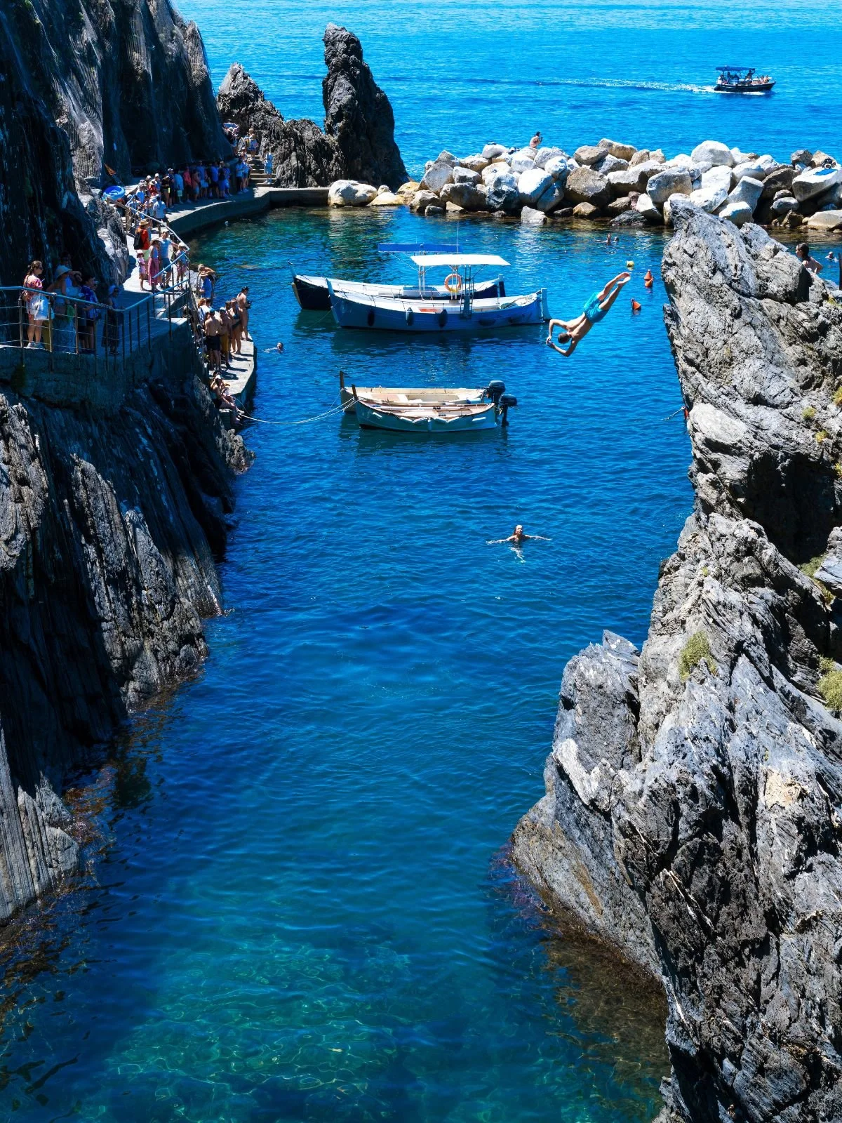 A scenic coastal scene featuring a rocky cove with boats anchored in clear blue water, people swimming, and a diving person mid-air diving into the water. A boat with a canopy floats near the rocks, and a boat with people is seen on the ocean in the background.