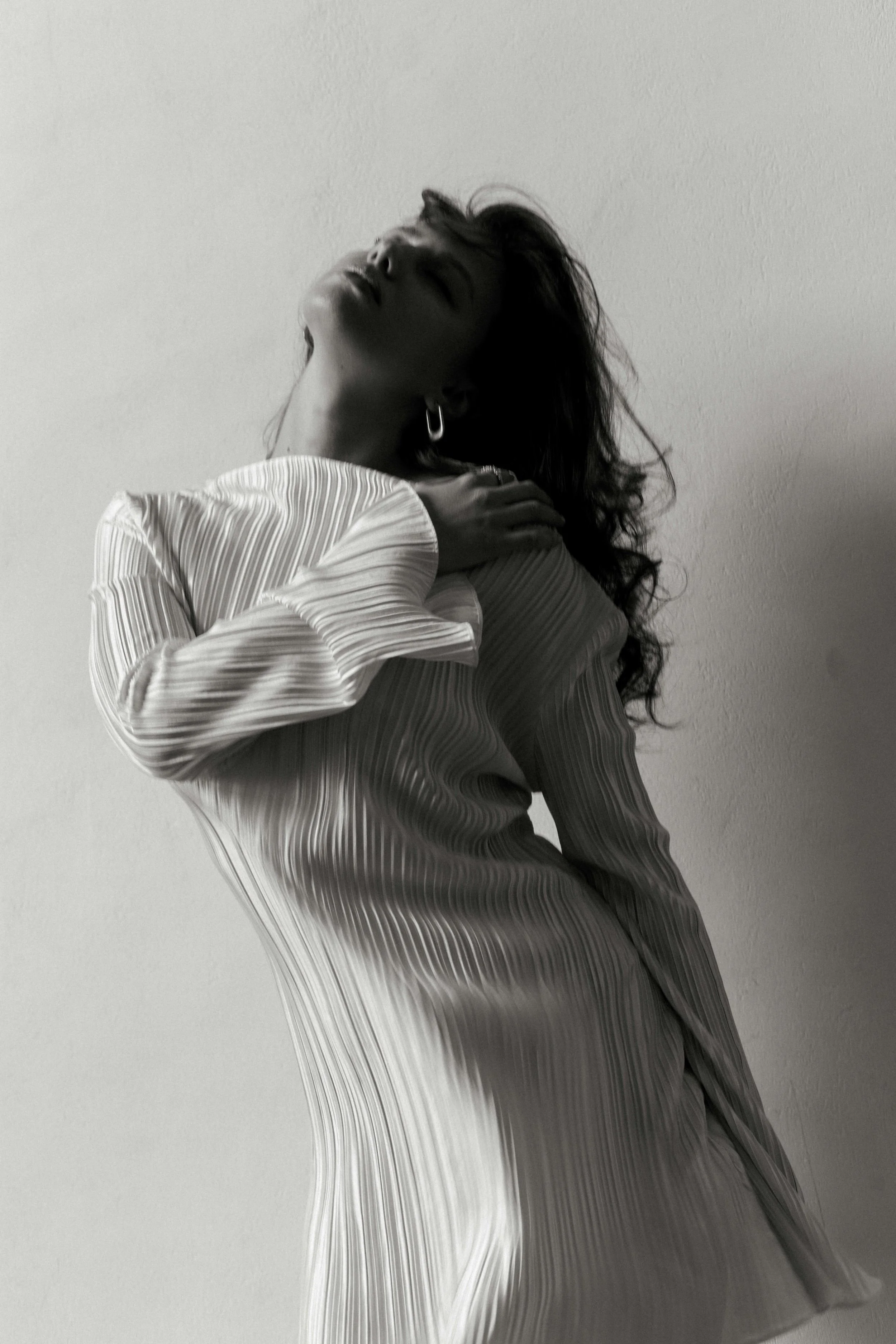 A woman in a pleated dress poses with her head tilted back, one hand on her neck, against a plain wall.