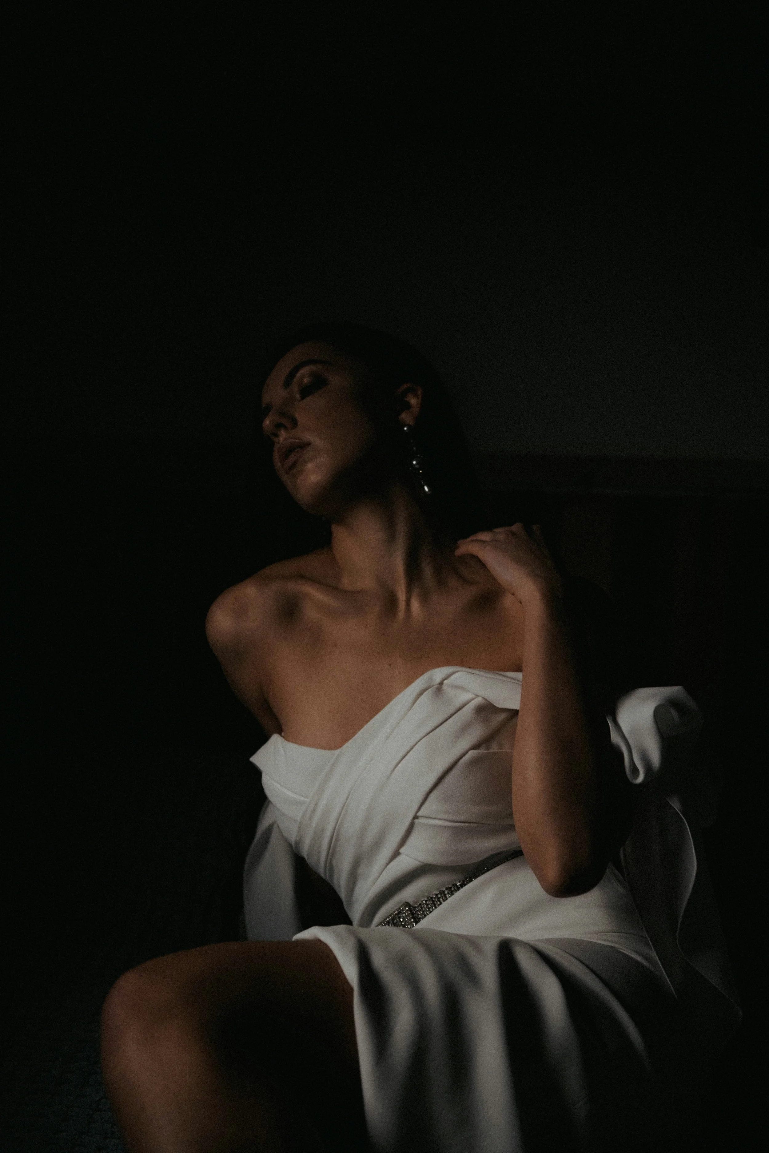 A woman in a white dress with an off-shoulder design and a side slit, sitting in a dimly lit space with soft lighting highlighting her face and shoulders.