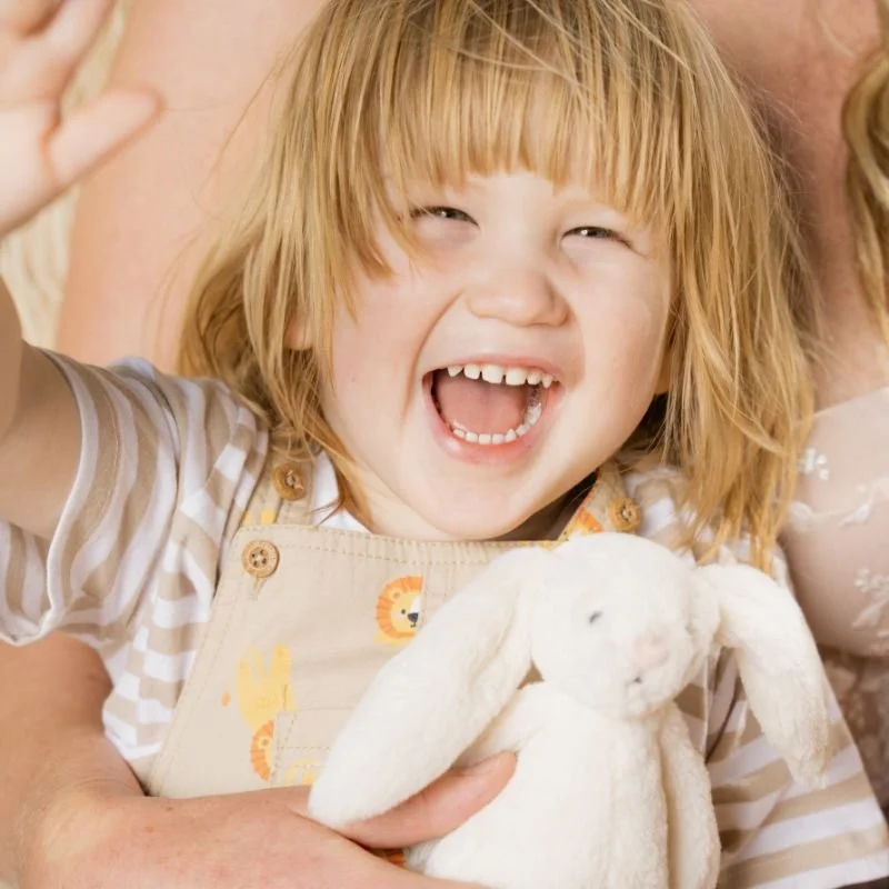 A young girl with red hair smiling and waving, holding a white stuffed bunny.