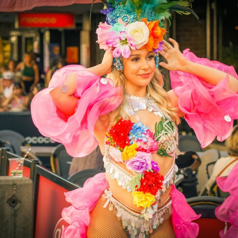 Woman dressed in vibrant, colorful costume with floral decorations and feathered headdress, performing or participating in a parade or festival.