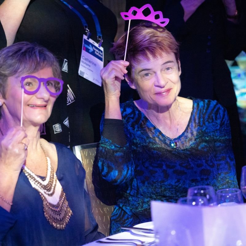 Two women smiling at a party or celebration, holding playful photo booth props including purple glasses and a tiara, sitting at a table with glasses and tableware.
