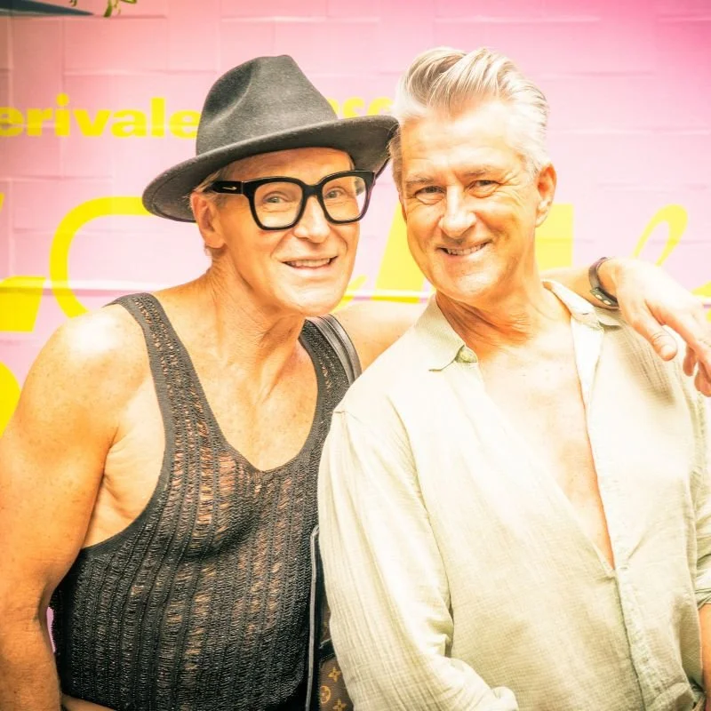 Two smiling men standing close together, one wearing a gray hat, black glasses, a black sleeveless top, and the other in a light-colored shirt with the top buttons open, against a pink background with yellow text.