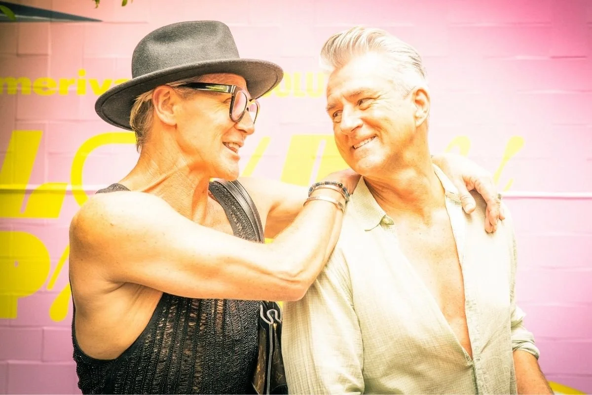 Two older adults, a woman and a man, are smiling and looking at each other. The woman is wearing a gray hat, glasses, and a black sleeveless top. The man is wearing a light-colored, partially unbuttoned shirt. They are standing close to each other, with the woman's arm around the man's shoulder, against a pink background with yellow writing.