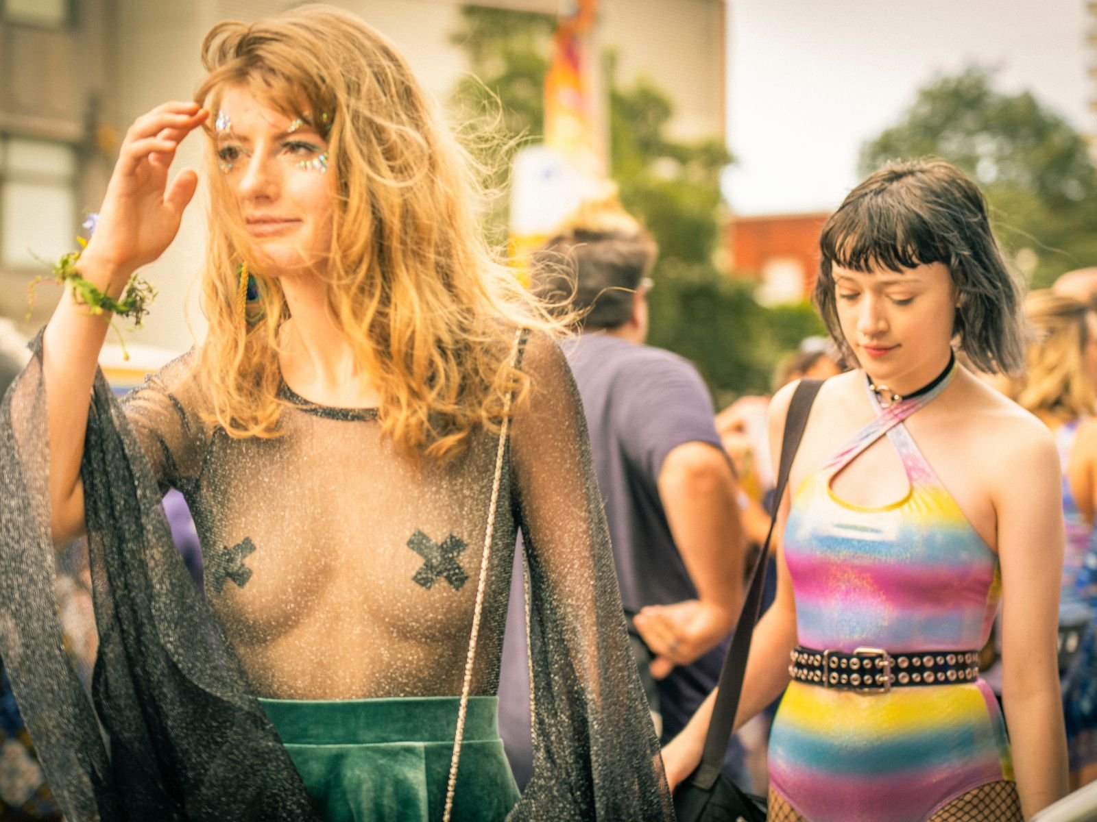 Two young women at an outdoor event, one with long, curly red hair and dusty pink lipstick, wearing a sheer black top with nipple covers and a green skirt, touching her hair and looking away, and the other with short, dark hair and bangs, wearing a colorful rainbow-patterned bodysuit with fishnet tights and a studded belt, looking down.